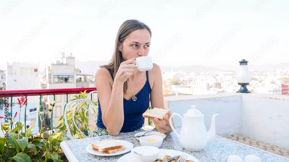Portrait of woman eating toast and drinking a tea for breakfast with pleasure in cafe on terrace with beautiful view on city. Front view.
