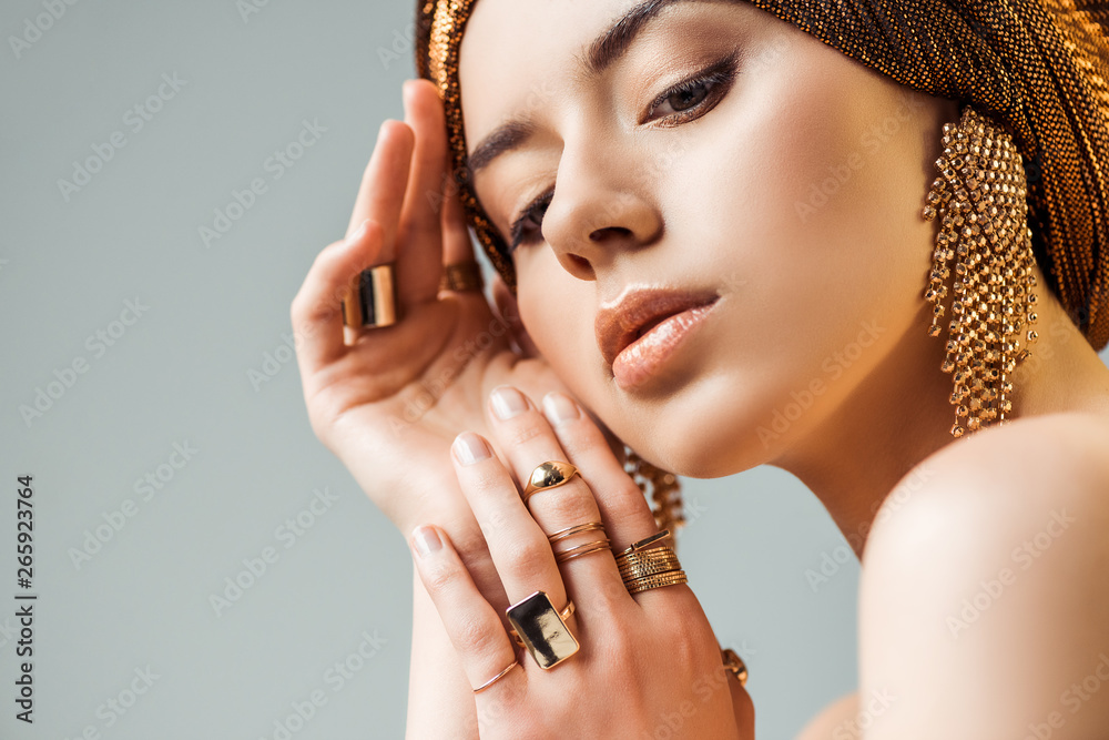 young tender naked woman with shiny makeup, golden rings and earrings in turban isolated on grey