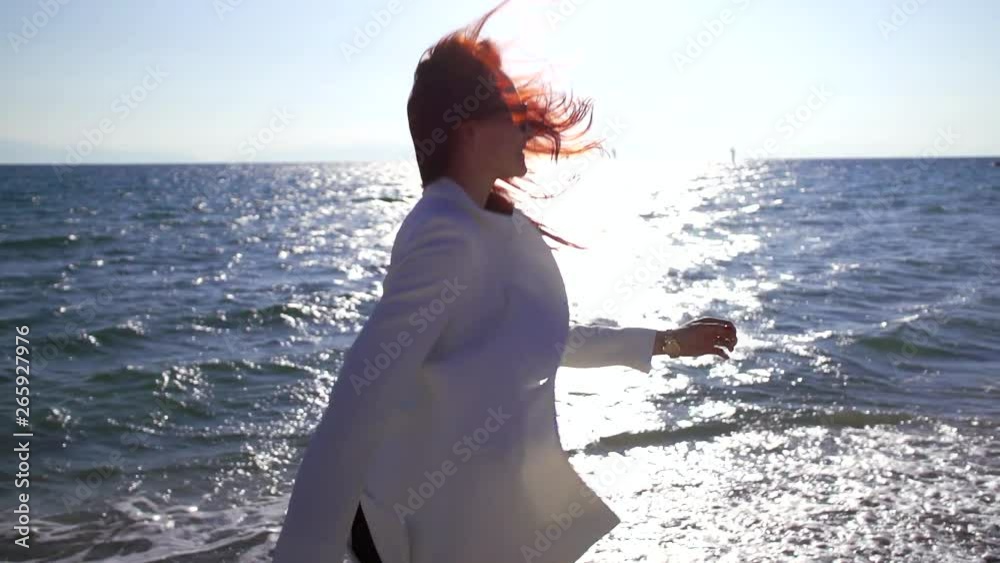 The girl in the white shirt and the sunglasses with bright hair jumps and spins on the background of the sea, is happy.
