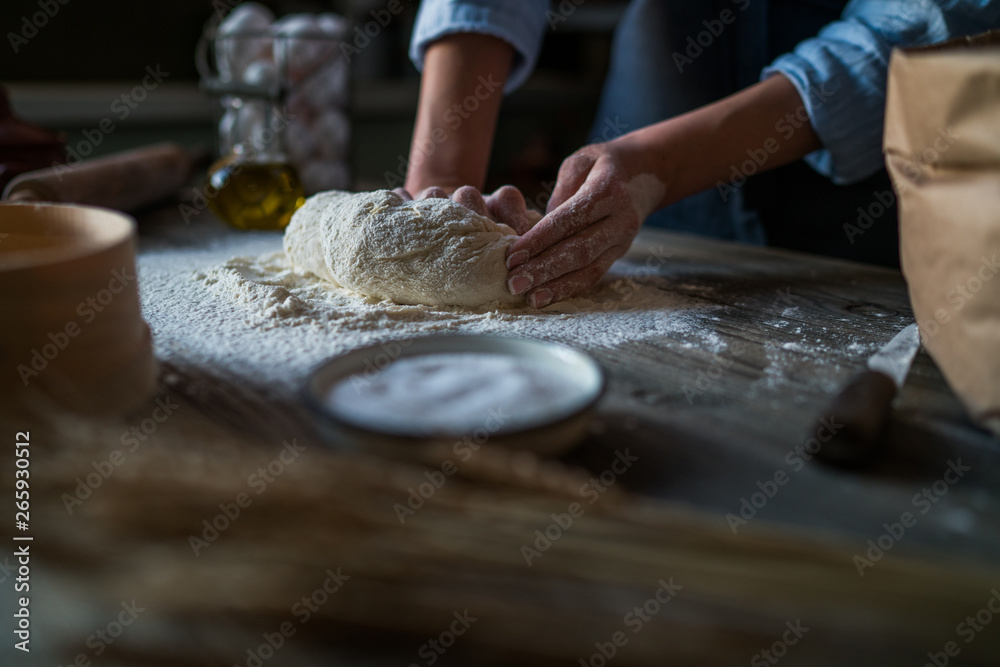 Beautiful women's hands knead the dough from which they will then make bread, pasta or pizza. Next to the kitchen utensils. The background is dark. Rustic style.