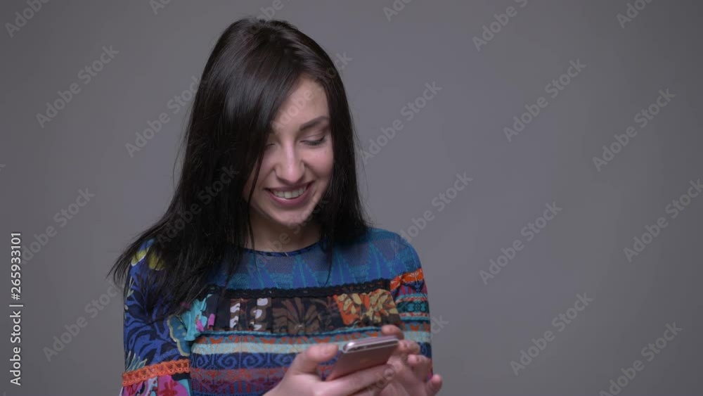 Closeup portrait of adult caucasian brunette female texting on the phone smiling and looking at camera with background isolated on gray Closeup portrait of adult caucasian brunette female texting on the phone smiling and looking at camera with background isolated on gray