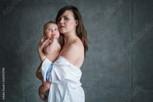 A young brunette mother in a white shirt, lowered on her shoulders, holds a shirt in the hands of a baby and gently presses it to her.