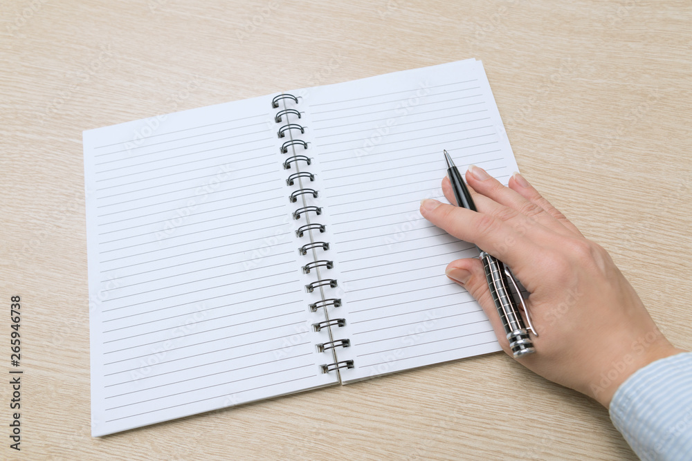 Top view of the table where there is a clean notebook. Women's hand hold a pen and prepare to write something. Top view of the table where there is a clean notebook. Women's hand hold a pen and prepare to write something.