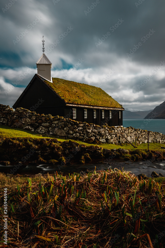 Long exposure of the small picturesque black wooden church of Funningur (Funnings Kirkja) with dramatic clouds and sun (Faroe Islands, Denmark, Europe)