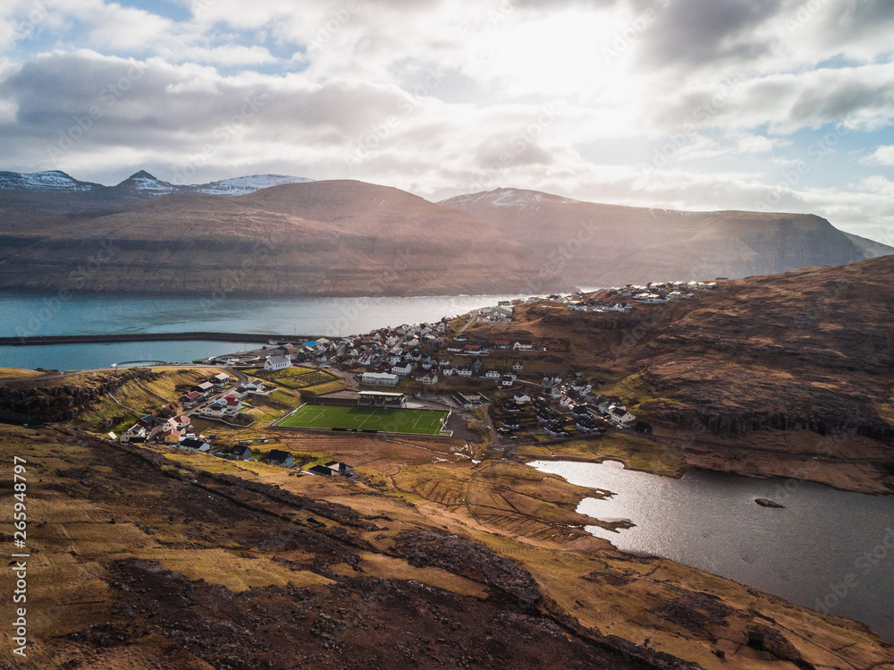 Aerial view of the village Eiði with snow-covered mountain range, football field and dramatic sky during sunset (Faroe Islands, Denmark, Europe)