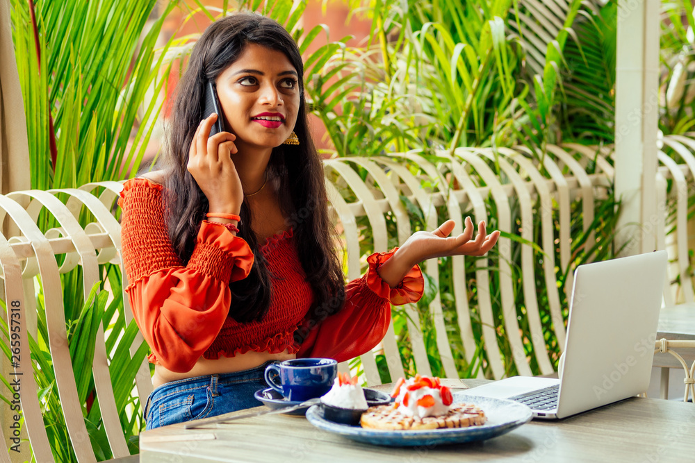 Indian businesswoman with laptop talking by phone in summer cafe . success student eat puncakes and drinking masala tea Indian businesswoman with laptop talking by phone in summer cafe . success student eat puncakes and drinking masala tea