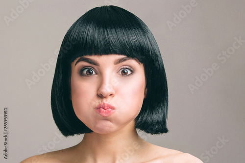 Young woman with black hair posing on camera. Expressive emotional model with bob haircut. Isolated on light background. Studio shot. Mouth full of air.