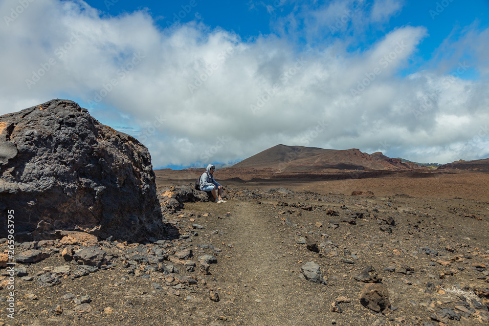 Tired traveler sitting near huge lava stone. Sunny day with beatiful fluffy clouds on the bright blue sky. Nationa Park Teide, Tenerife, Spain