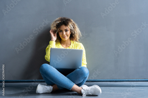 Quadro su tela Happy young woman sitting on the floor with crossed legs and using laptop on gray background