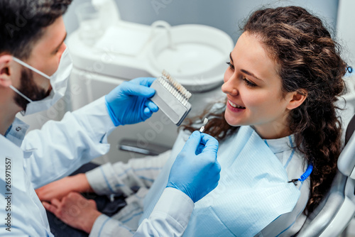 Close up portrait of charming young woman sitting in dental chair while stomatologist selecting color tone of her teeth.