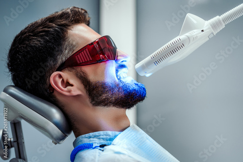 Unrecognizable dentist whitening teeth of young man sitting in dental chair in modern clinic. Side view. photo