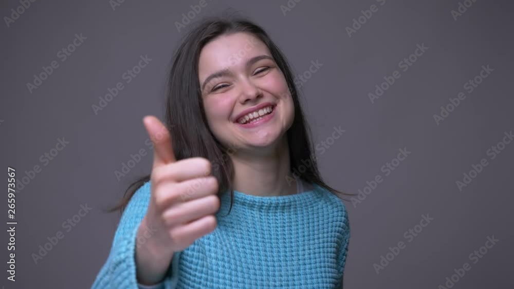 Closeup shoot of young pretty brunette female gesturing a thumb up and smiling happily looking at camera with background isolated on gray