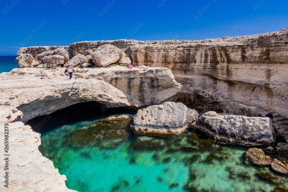 Archaeological site and tourist resort of Roca Vecchia, Puglia, Salento, Italy. Turquoise sea, clear blue sky, rocks, sun, in summer. The Cave of Poetry. Tourists take pictures and sunbathe.