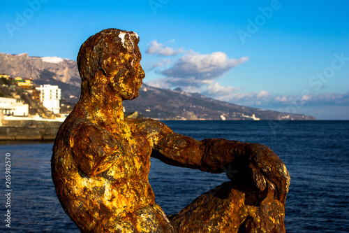A rusty brown statue of a man falling apart and abandoned against the backdrop of nature, the sea and the mountains. Metal sculpture the whole covered with rust.