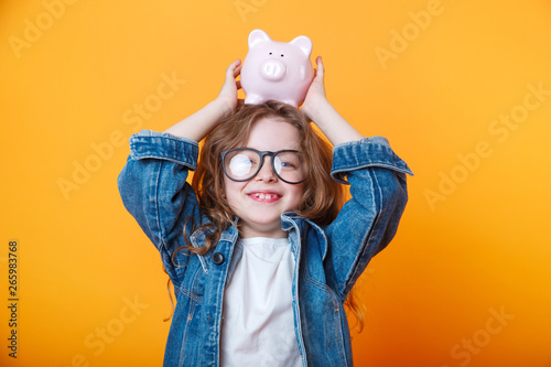 Cute little girl in eyeglasses shaking piggy box on orange background