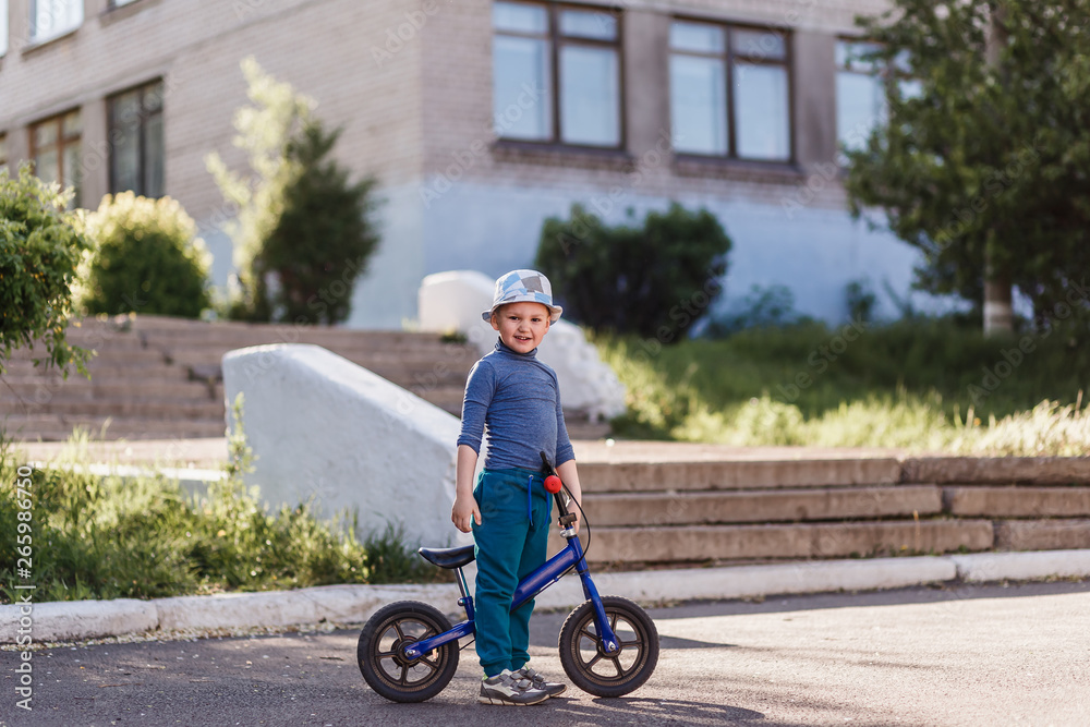 a four-year-old smiling boy in a hat sits on a blue running bike. Bicycle without pedals