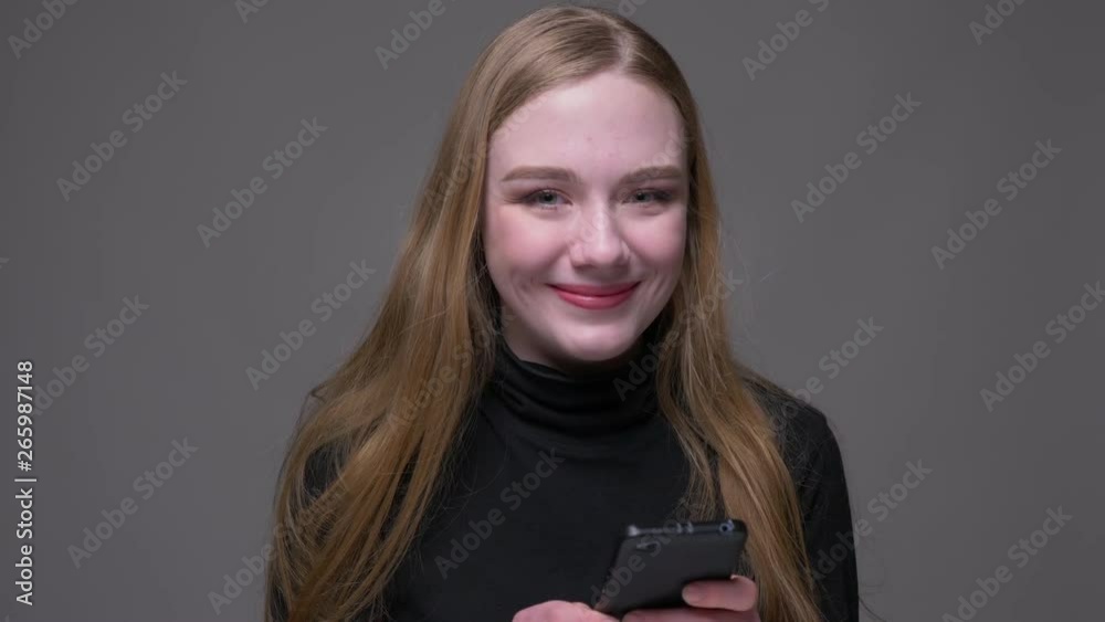 Closeup portrait of young attractive brunette female using the phone and smiling happily looking at camera with background isolated on gray Closeup portrait of young attractive brunette female using the phone and smiling happily looking at camera with background isolated on gray