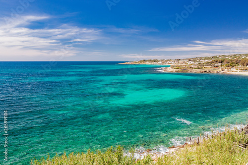 Spectacular view of the Apulian coast in Leuca, in Puglia, Salento, Italy. Turquoise sea, clear blue sky, rocks, sun, wind, white clouds, lush vegetation in summer. Populated area with white houses.