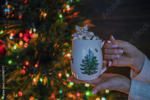 Woman drinking hot tea or coffee from festive cup. Girl enjoying winter morning or evening. Christmas Holidays. Christmas tree with decorations and lights in the room. Hot drinks and people concept.