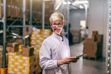 Young Caucasian blonde woman in white uniform using tablet in warehouse.Young Caucasian blonde woman in white uniform using tablet in warehouse.