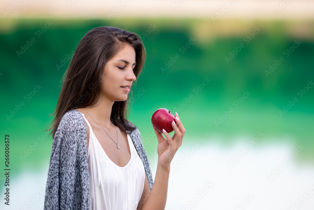 Young beautiful girl in white dress eating ripe organic apple. She is on the shore of the lake and enjoy a picnic and outdoor recreation. Young beautiful girl in white dress eating ripe organic apple. She is on the shore of the lake and enjoy a picnic and outdoor recreation.