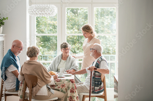 Group of senior friends with helpful carer sitting together at the table at nursing home dining room