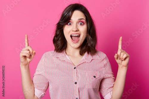 Srudio shot of young brunette girl posing isolated over pink background, points fingers up, having great, female wears striped shirt, has wavy hair, stands with opened mouth. People emotions concept. photo