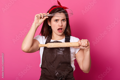 Studio shot of pleasant looking female with shocked facial expression, wears white t shirt, brown apron, head haed band, holding whisk and rolling pin, standing over pink wall with empty space.