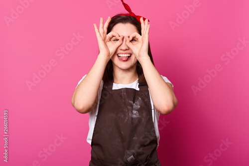 Studio shot of cheerful smiling young woman, covering her eyes with ok signs, expresses joy, model poses against pink background, being glad and joyful. People, body language and gesture concept. photo