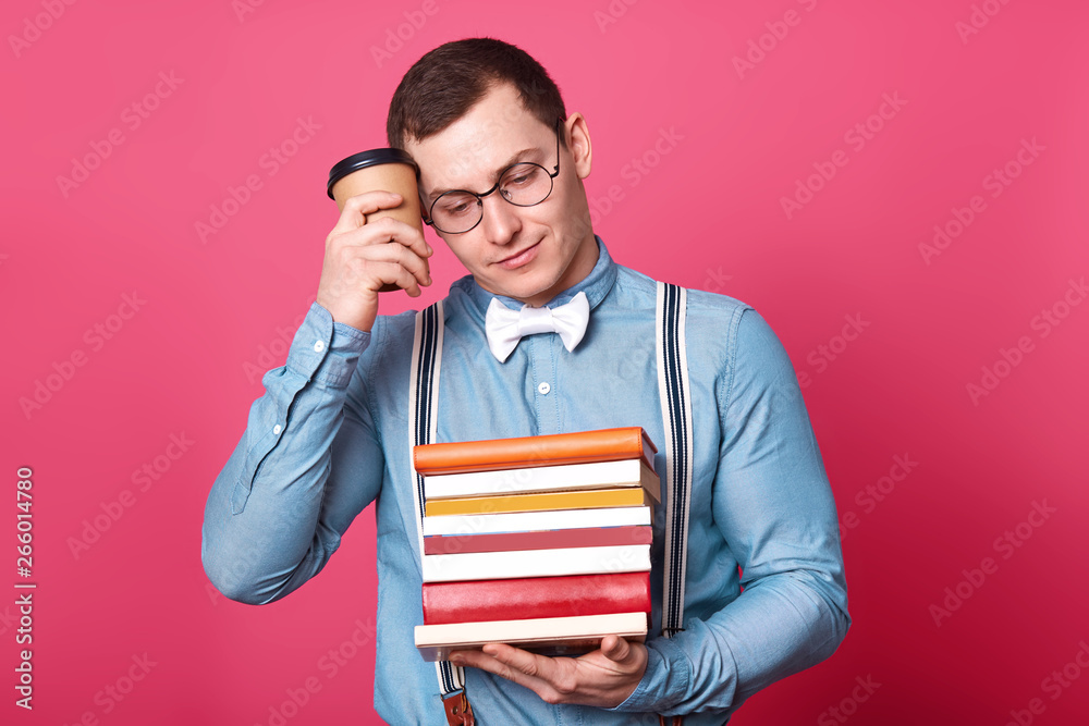 Exhausted athletic student touches his temple with papercup of drink, looks aside, holds books in one hand, having lunch during woring day, looks stressful, isolated over pink background in studio. Exhausted athletic student touches his temple with papercup of drink, looks aside, holds books in one hand, having lunch during woring day, looks stressful, isolated over pink background in studio.
