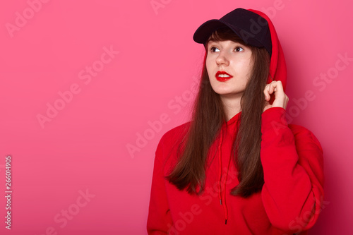Close up portrait of young cute brunette teenager girl in casual red hoodie and cap, posing with bright listick on pink background looking aside at copy space. Advertising and people concept.