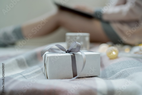 Christmas online shopping. Close up white christmas gift box with pink ribbon, on the blurred cozy home background. Woman buys presents, prepare to xmas eve. Cozy blanket. Toned image.