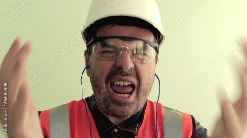 Caucasian unshaven man worker dressed in a reflective vest, protective glasses and white helmet nervously arguing, shouting, waving his arms to the camera. Isolated on light green background