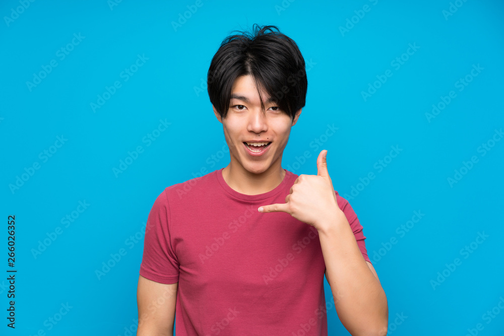 Asian man with red shirt over isolated blue wall making phone gesture Asian man with red shirt over isolated blue wall making phone gesture
