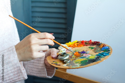 Close-up of female hands in a pink sweater holding a brush and a palette with multi-colored acrylic paints against a white canvas