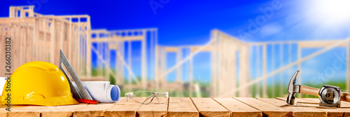 Construction Tools On Wooden Table With New House Framework In Background - Housing Industry Concept 
