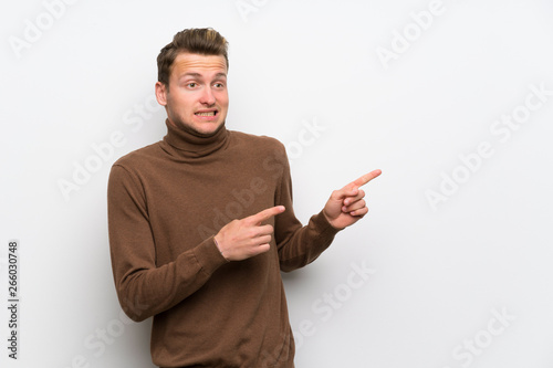 Blonde man over isolated white wall frightened and pointing to the side