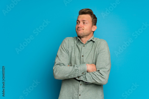 Blonde man over isolated blue wall making doubts gesture while lifting the shoulders