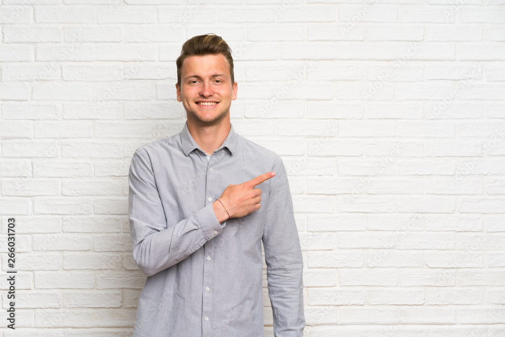 Blonde man over white brick wall pointing to the side to present a product Blonde man over white brick wall pointing to the side to present a product