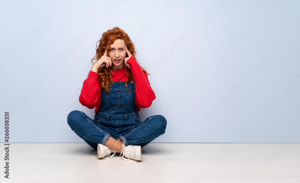 Redhead woman with overalls sitting on the floor having doubts and thinking Redhead woman with overalls sitting on the floor having doubts and thinking
