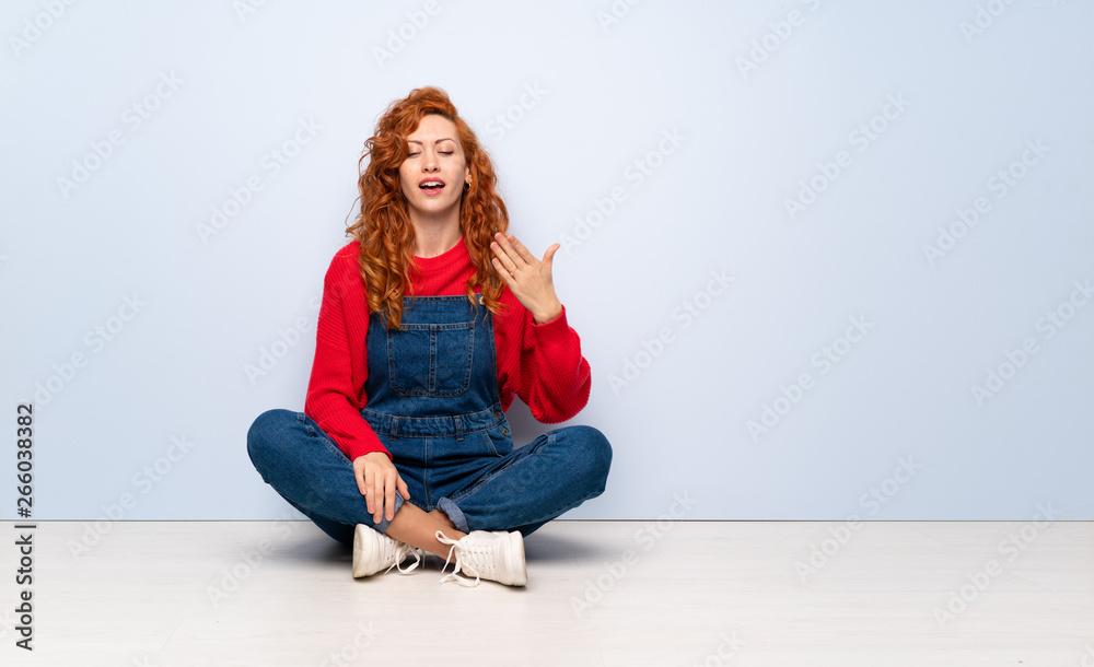 Redhead woman with overalls sitting on the floor with tired and sick expression