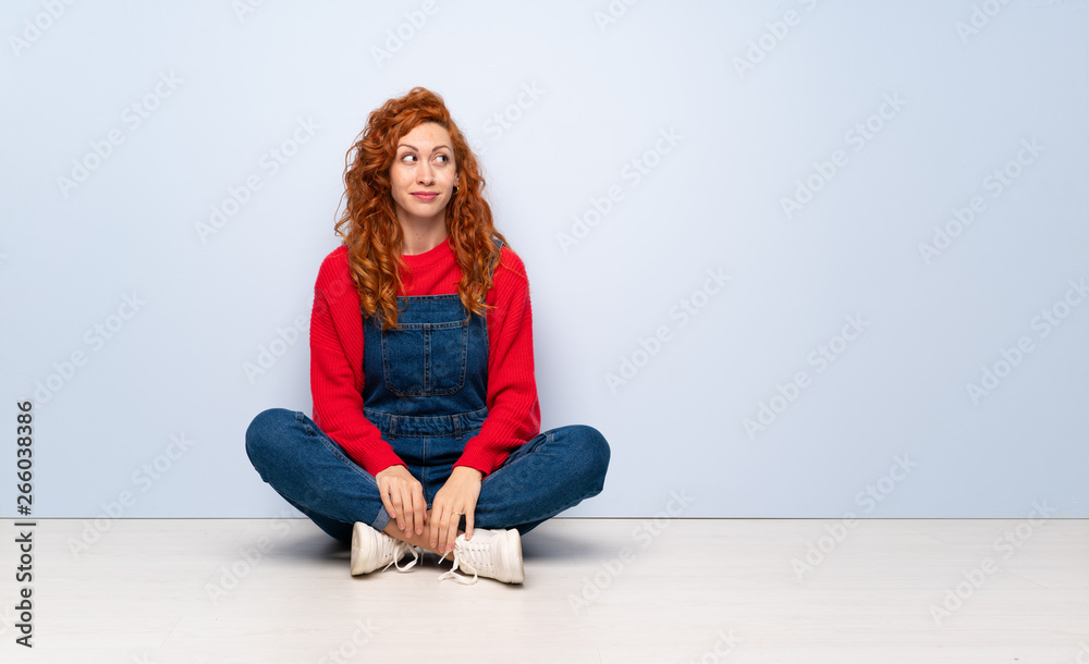Redhead woman with overalls sitting on the floor making doubts gesture looking side Redhead woman with overalls sitting on the floor making doubts gesture looking side