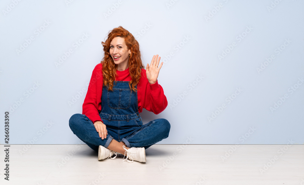 Redhead woman with overalls sitting on the floor saluting with hand with happy expression Redhead woman with overalls sitting on the floor saluting with hand with happy expression