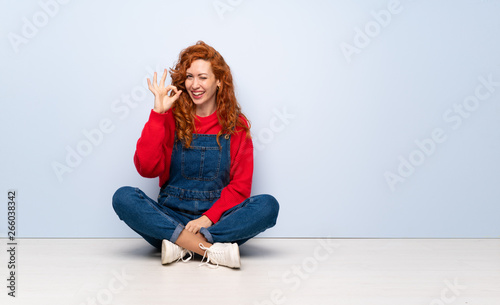 Redhead woman with overalls sitting on the floor showing ok sign with fingers