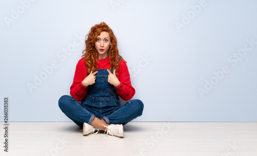 Redhead woman with overalls sitting on the floor with surprise facial expression