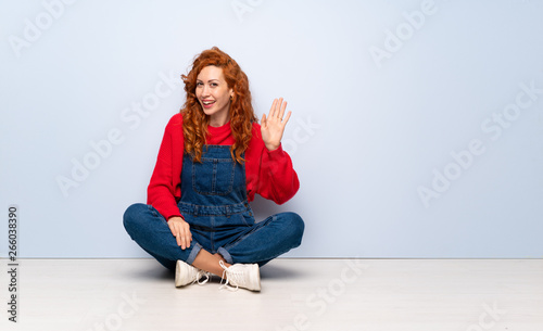 Redhead woman with overalls sitting on the floor saluting with hand with happy expression