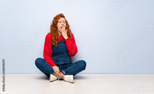 Redhead woman with overalls sitting on the floor yawning and covering wide open mouth with hand