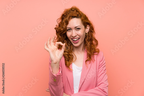 Redhead woman in suit over isolated pink wall showing an ok sign with fingers