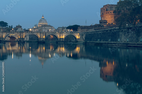 River Tiber in Rome and St Peters Cathedral at night. Reflections in the water of illuminated buildings. Aurelius Bridge over river with Castel Sant'Angelo at dusk