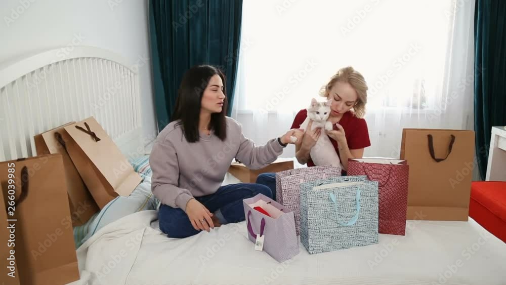 Two attractive women sit on bed and discuss new clothes from shopping. Blogging concept, recording video, friendship and consumerism concept